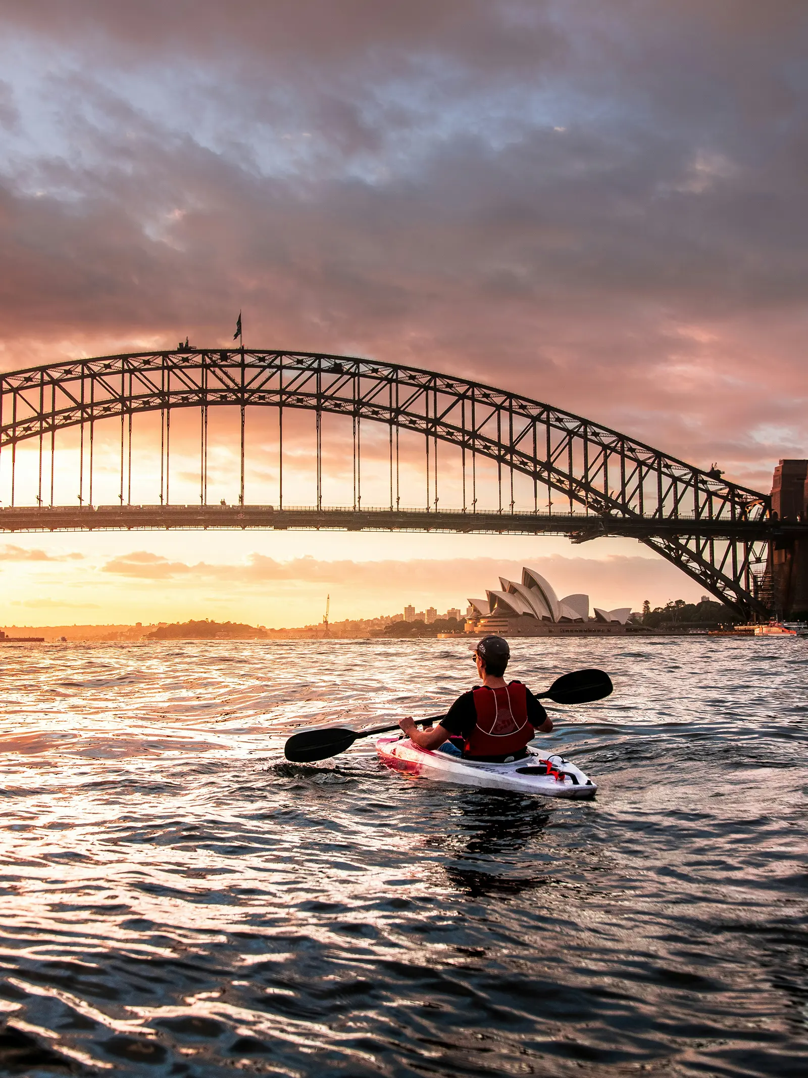 A kayaker paddling across Sydney Harbour at sunset, with the Harbour Bridge and Opera House behind.